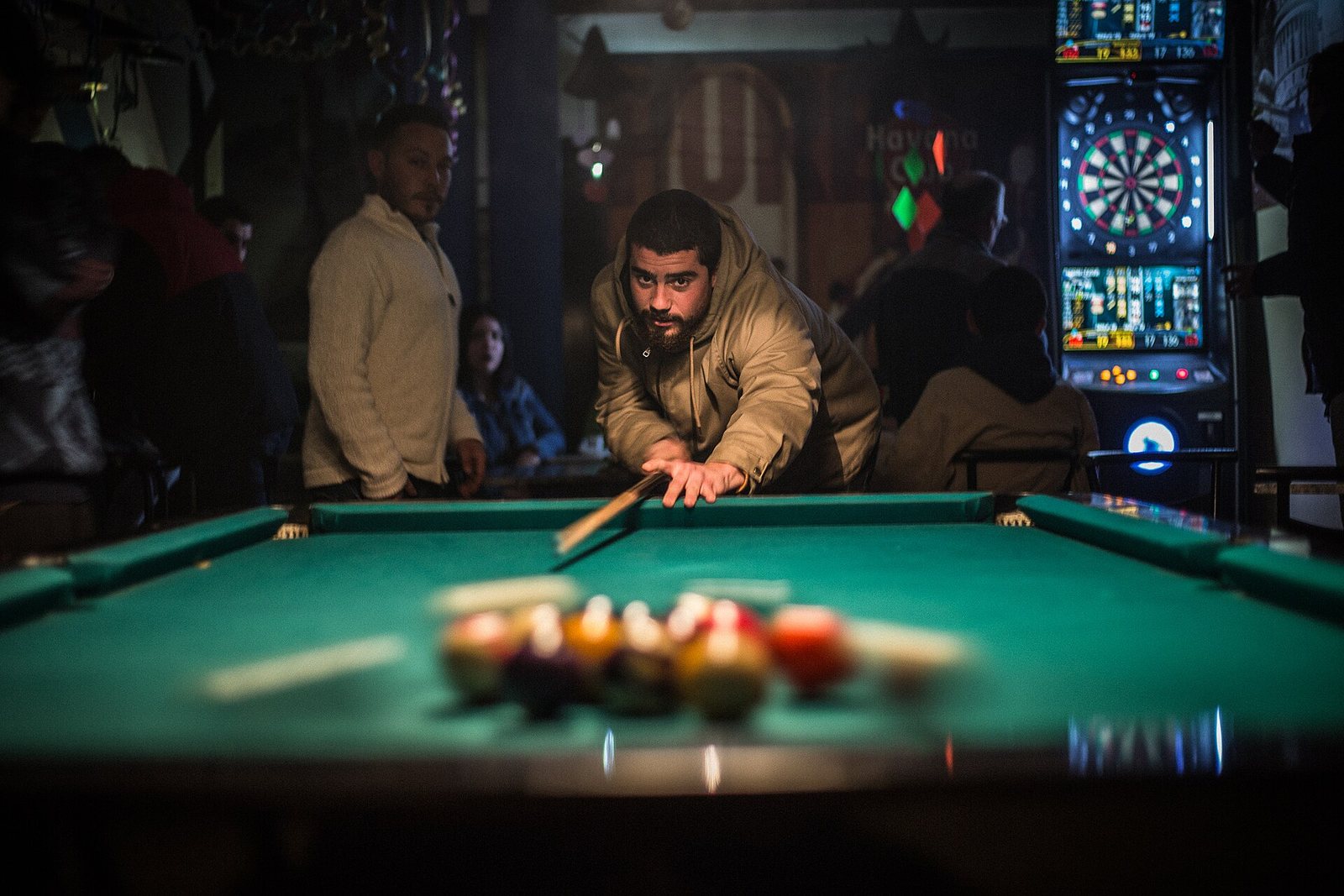 A player leans over a green-felt pool table lining up the break in a dim, neon-lit hall, the racked balls blurred in the foreground.